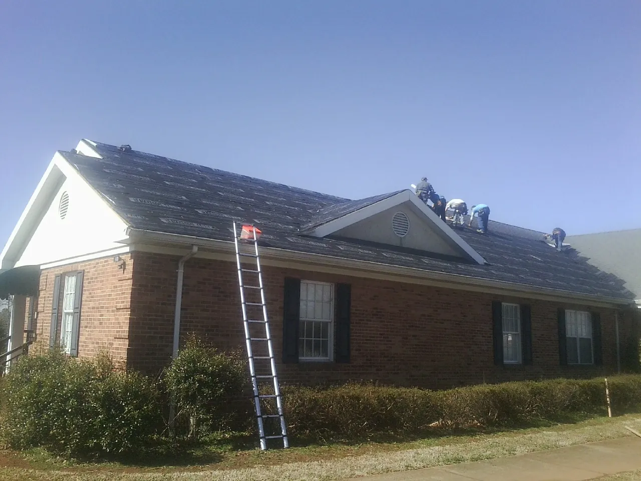 Crew tearing shingles off a brick ranch roof with a ladder set — Metal Roof Repair work by our crew in Rochelle
